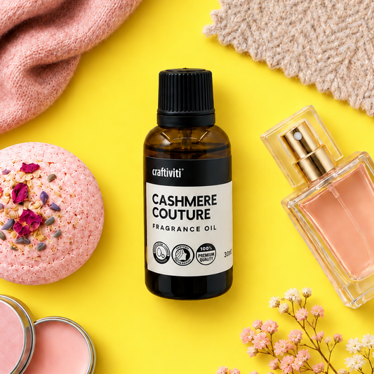 A bottle of cativiti Cashmere Couture Fragrance Oil is styled in a flat-lay arrangement on a bright yellow background, surrounded by a pink bath bomb, a pink lip balm tin, a clear perfume bottle, and soft pink fabrics.