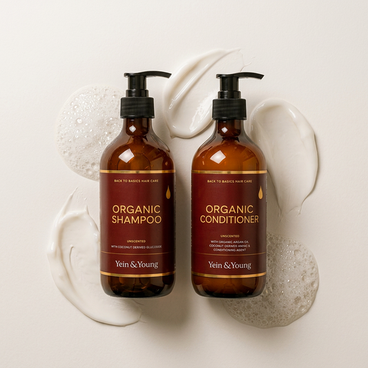 Two amber glass pump bottles of Yein & Young Organic Shampoo and Organic Conditioner displayed on a marble bathroom counter with a sink, candle, and greenery in the background.