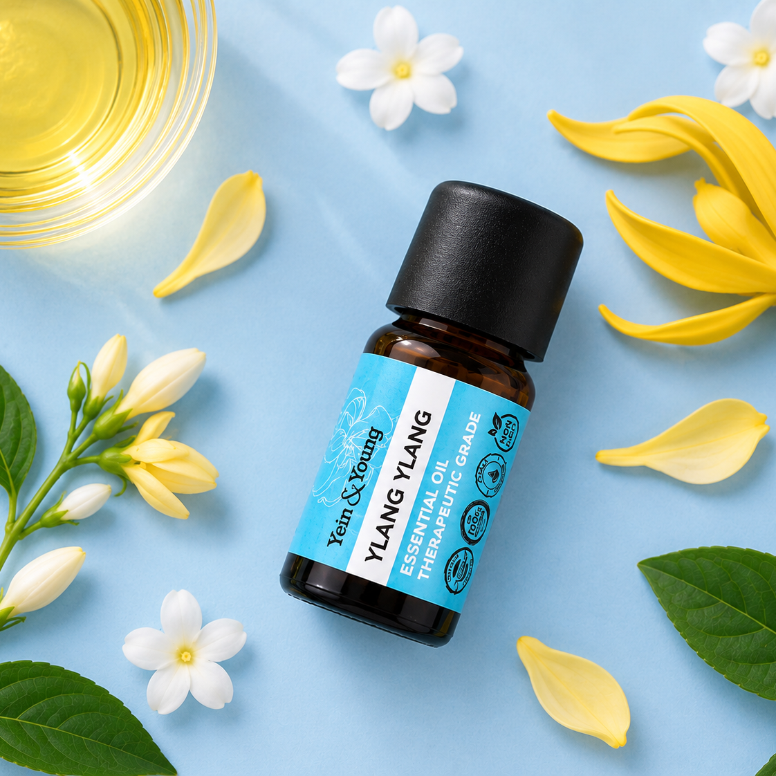 A bottle of Ylang Ylang essential oil oil by Yein & Yang, styled in a flat-lay with yellow flowers, white blossoms, green leaves, and a glass of golden oil on a light blue background.